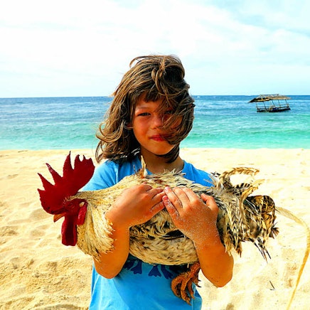 Ein Junge steht mit einem Hahn auf dem Arm am Strand.