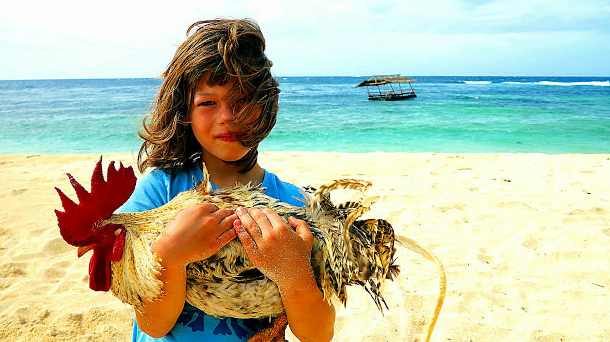 Ein Junge steht mit einem Hahn auf dem Arm am Strand.