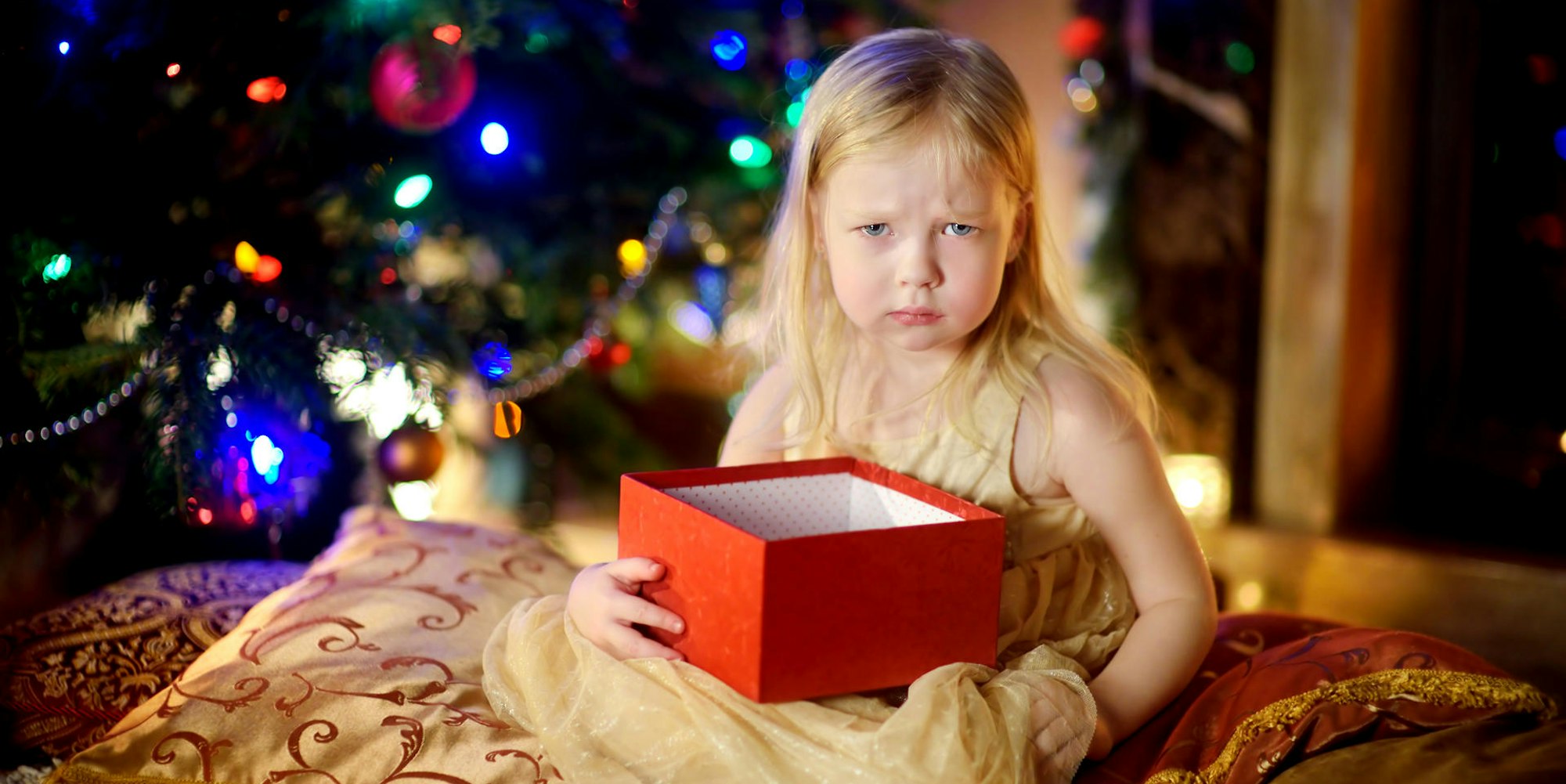 Ein Kind hält ein Weihnachtsgeschenk und sitzt vor dem Tannenbaum, es schaut schlecht gelaunt.