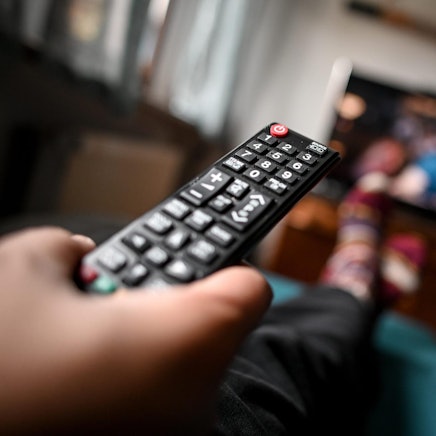 Eine Person hält eine Fernbedienung in der Hand, im Hintergrund ist unscharf ein Fernsehgerät zu sehen.
