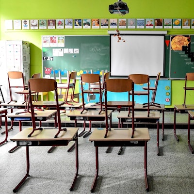 In einem leeren Klassenzimmer stehen Stühle auf Tischen. Vorne ist eine Tafel zu sehen, vor der Tafel ist eine Leinwand.
