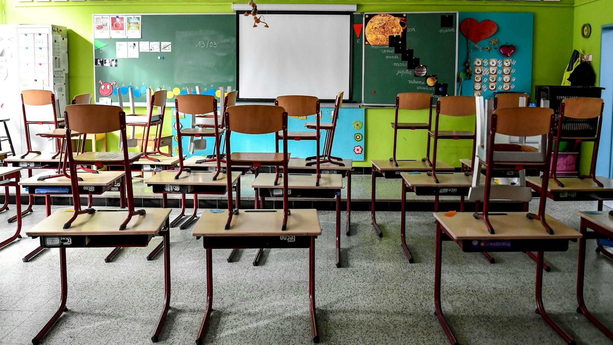 In einem leeren Klassenzimmer stehen Stühle auf Tischen. Vorne ist eine Tafel zu sehen, vor der Tafel ist eine Leinwand.