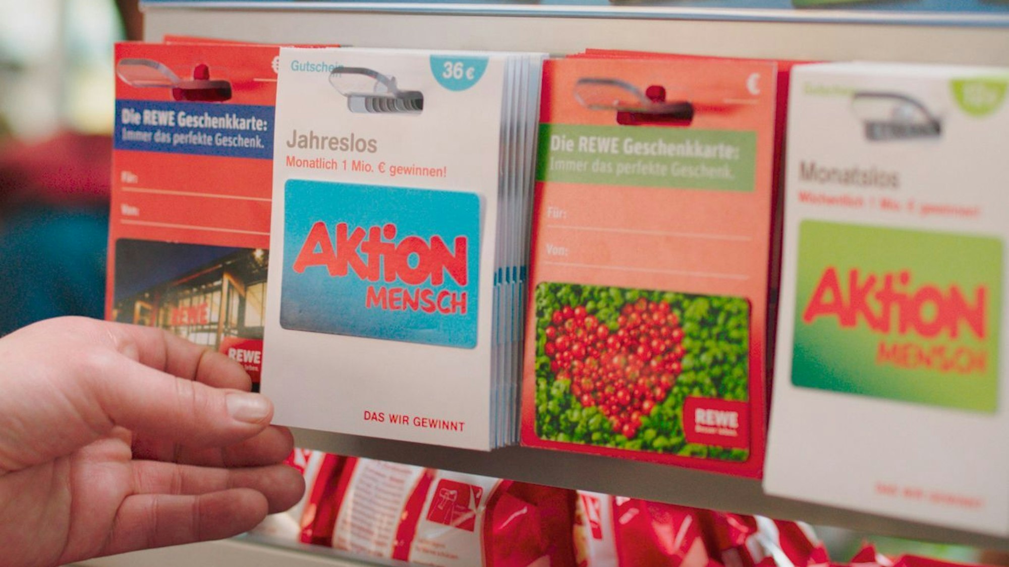 Ein Los der Aktion Mensch wird von einer Frauenhand aus einem Regal bei Rewe genommen. Daneben befinden sich weitere Lose.