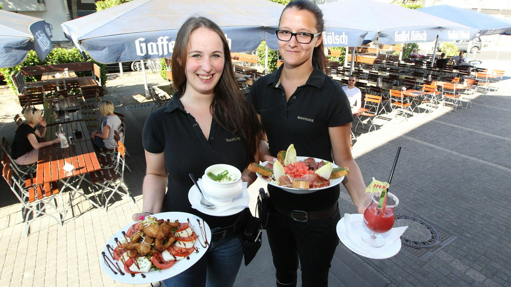 Zwei Frauen halten vier Gerichte in den Hönden. Im Hintergrund stehen Tische und Stühle unter Sonnenschirmen.