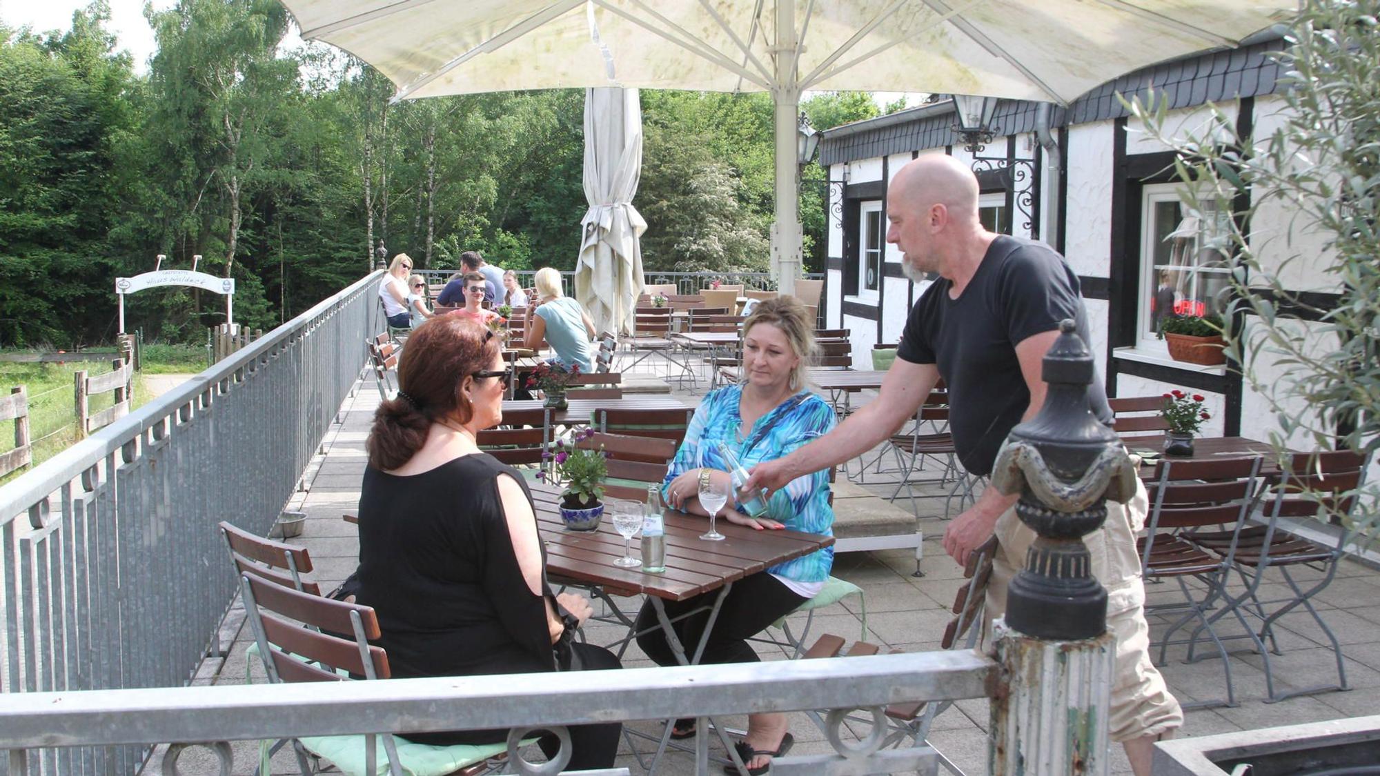 Zwei Frauen sitzen an einem Tisch auf einer Außenterrasse. Ein Mann bedient die beiden.