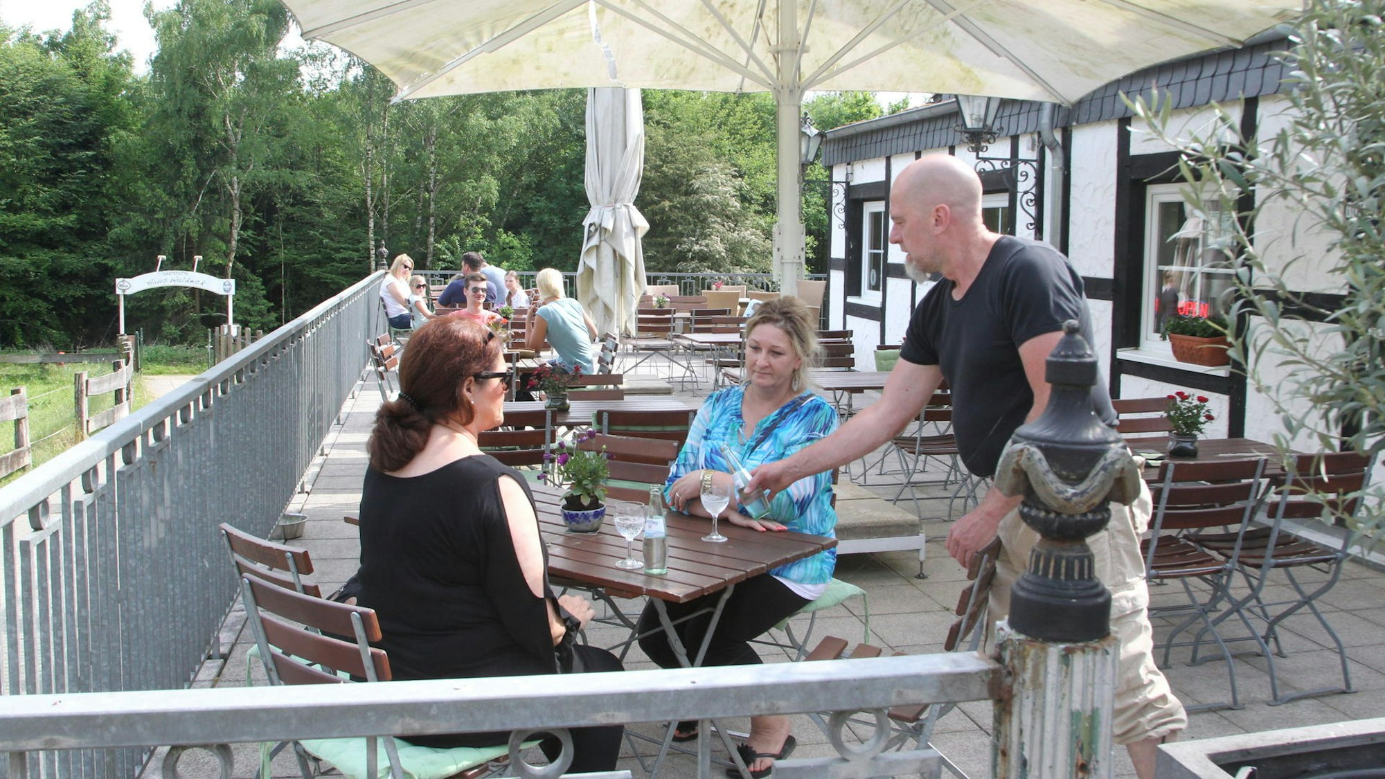 Zwei Frauen sitzen an einem Tisch auf einer Außenterrasse. Ein Mann bedient die beiden.