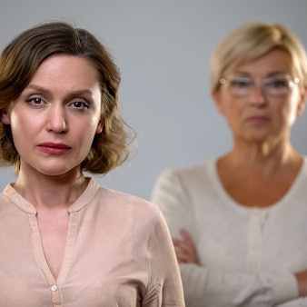 Eine Frau mittleren Alters schaut in die Kamera, im Hintergrund ist unscharf ihre Mutter zu sehen.