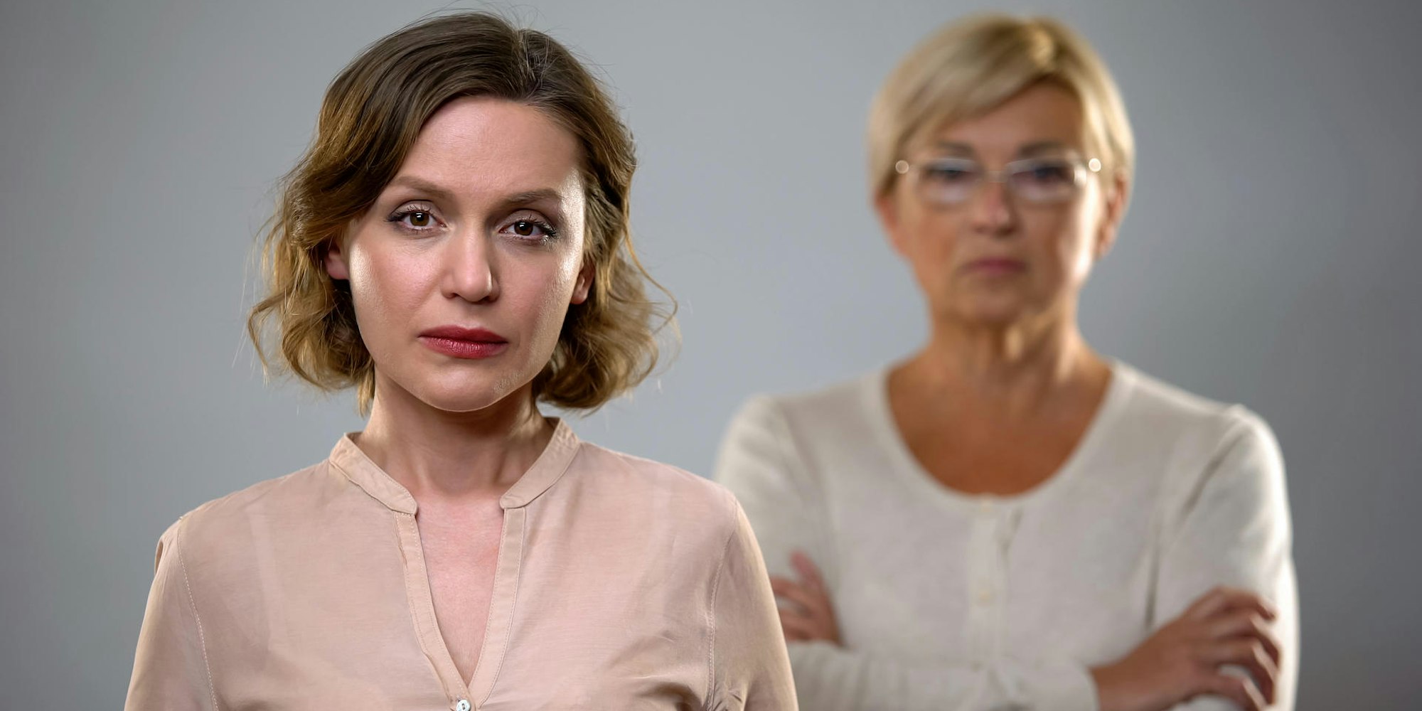 Eine Frau mittleren Alters schaut in die Kamera, im Hintergrund ist unscharf ihre Mutter zu sehen.