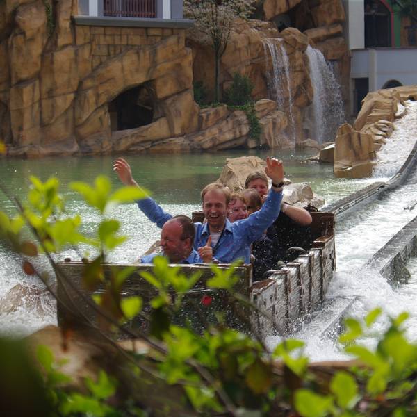 Besucher sitzen in einem Fahrgeschäft im Phantasialand, das durch eine Wasser-Fläche fährt. Ein Mann hat die Arme gehoben.