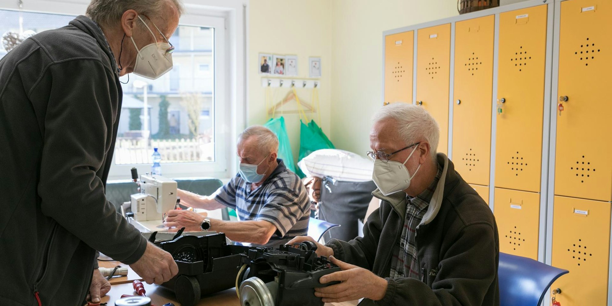 Ein Mann sitzt an einem alten Gerät und begutachtet es. Im Hintergrund sitzt ein Mann an einer Nähmaschine.