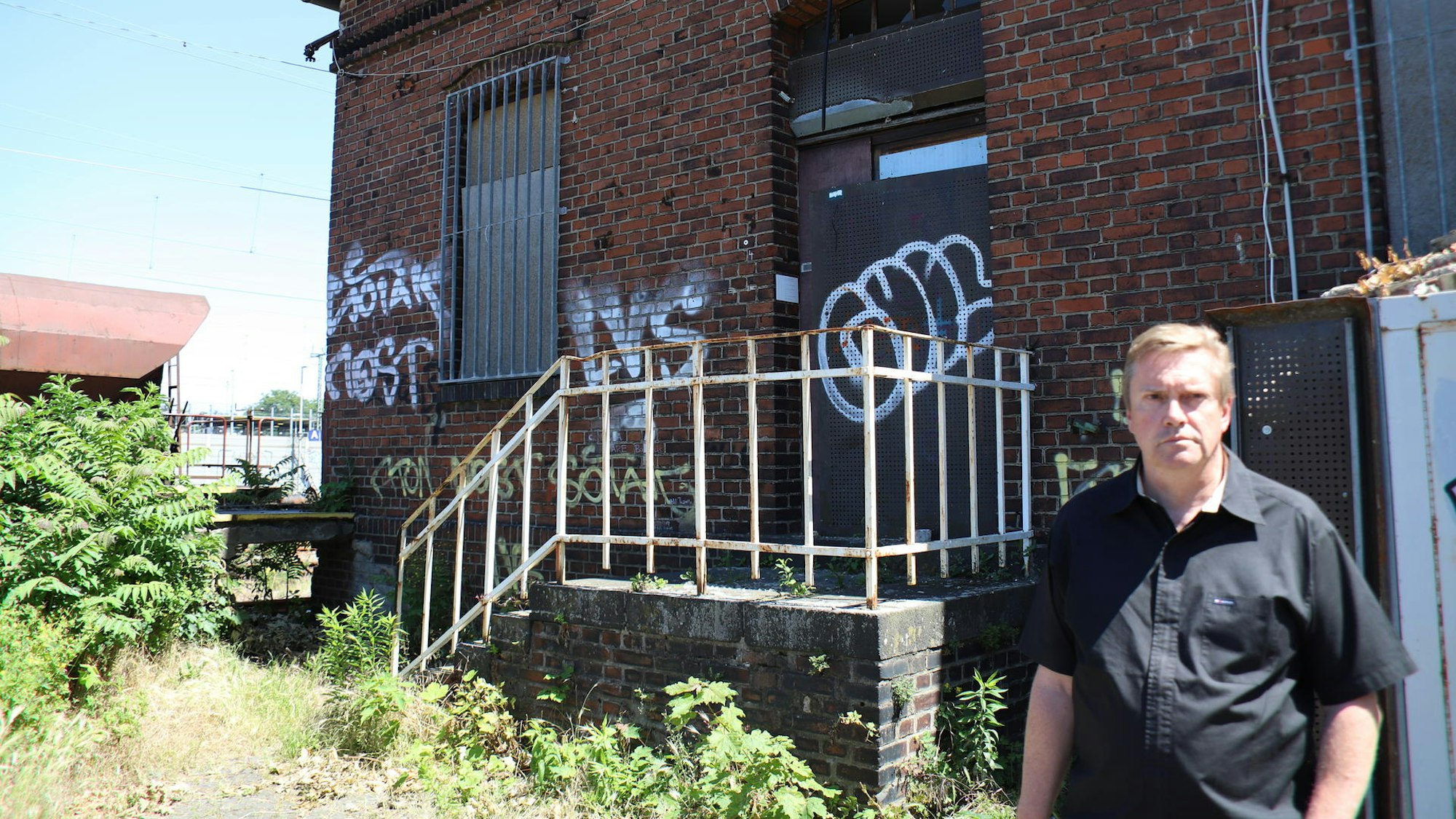 Ein Mann in einem schwarzen Hemd steht vor dem alten Lagerhaus. Im Hintergrund steht ein Güterwagen der Bahn.