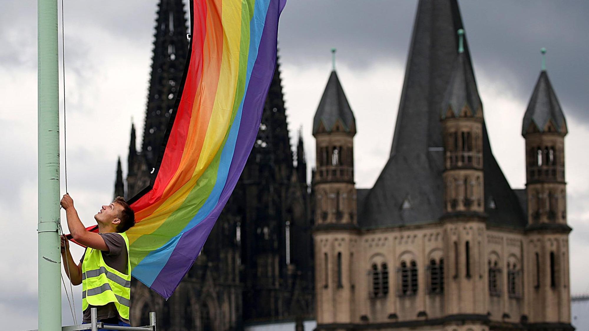 Ein Mann hisst eine Regenbogenfahne, im Hintergrund ist eine Kirche zu sehen.
